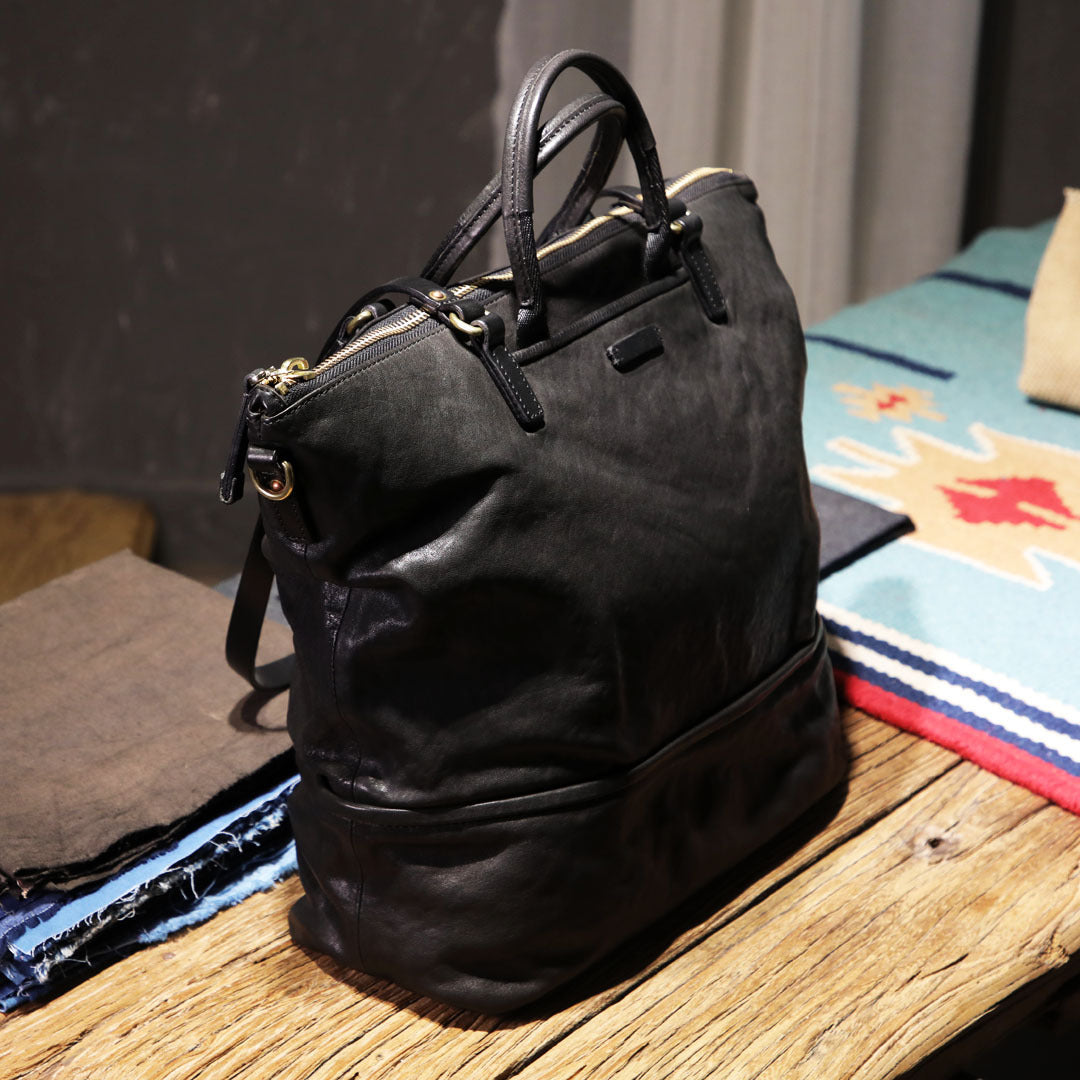 Closed black leather tote bag placed on a wooden table surrounded by vibrant Latin American fabrics.