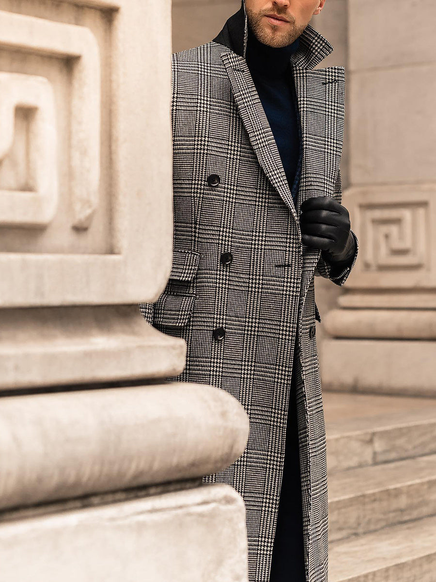 Sophisticated man wearing a gray checkered overcoat, leaning against a Roman-style building while adjusting his sleeve or cufflinks.